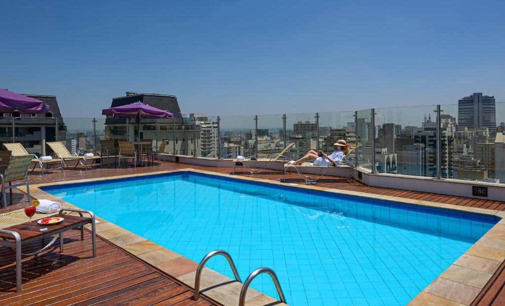 Piscina azul com piso de madeira, com algumas cadeiras e guarda-sol com vista para a cidade de prédios durante o dia, ilustrando post Hotéis Mercure em São Paulo.