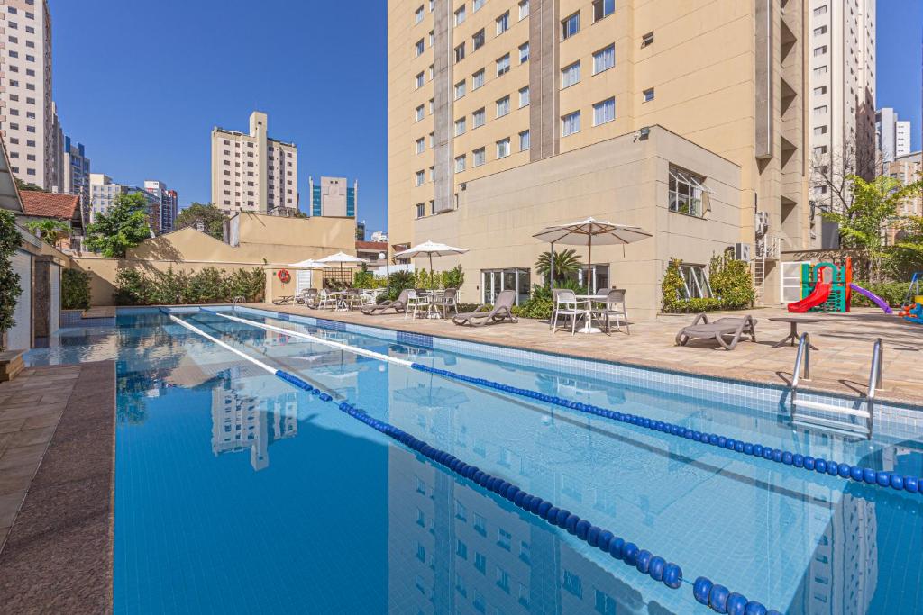 Parte de fora do hotel com piscina grande e azul, com algumas cadeiras e mesa com guarda-sol e ao fundo outros prédios, ilustrando post Hotéis Mercure em São Paulo.