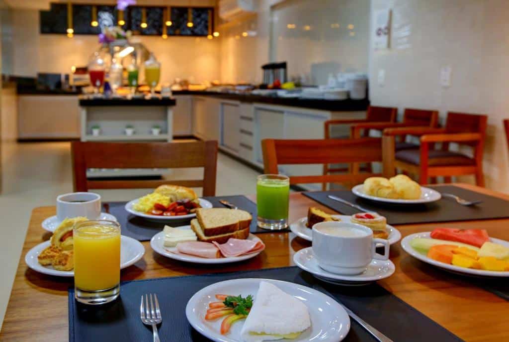 Mesa com café da manhã de hotel com frutas, pães, tapioca, sucos e xícara de café.
