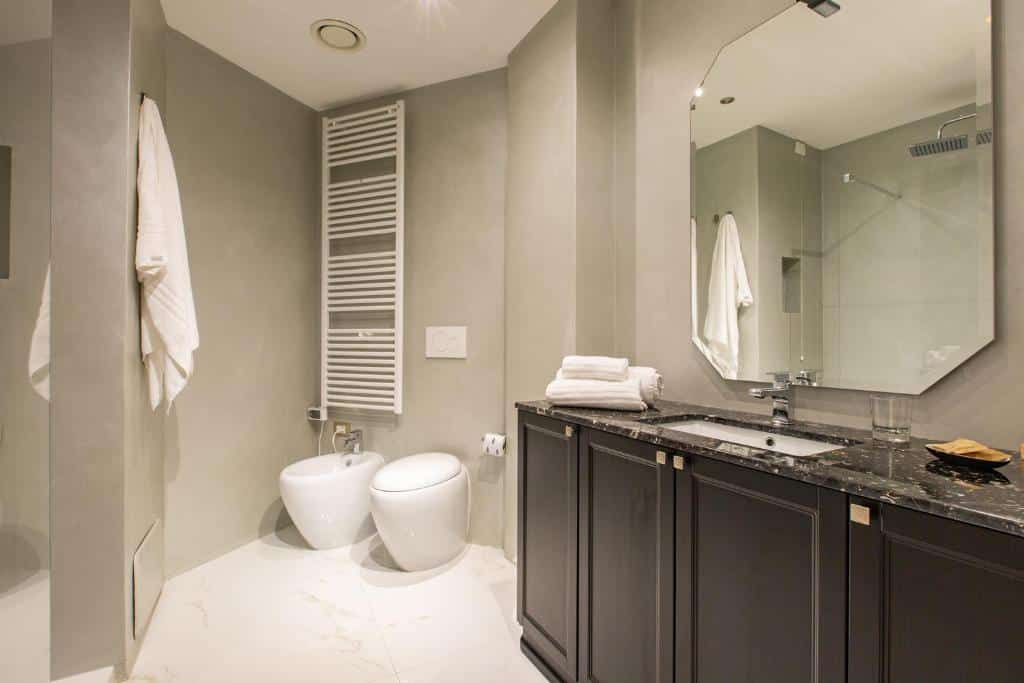 Banheiro do Missori Suites com um box, um bidê e um vaso sanitário, há um móvel de madeira preta onde está a pia, há um espelho amplo e toalhas brancas disponíveis