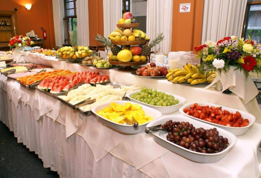 Mesa com várias frutas, como banana, uva, morango, melancia, abacaxi, entre outras e algumas flores na toalha branca, ilustrando post Hotéis perto da 25 de Março em SP.