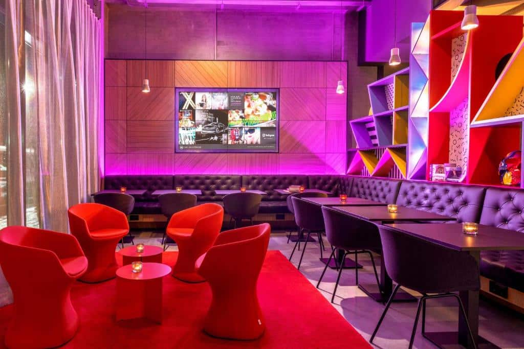 Lounge compartilhado do Moxy Milan Linate Airport com poltronas vermelhas, sofás estofadas roxos, mesas quadradas e cadeiras roxas, a iluminação mescla amarelo e rosa