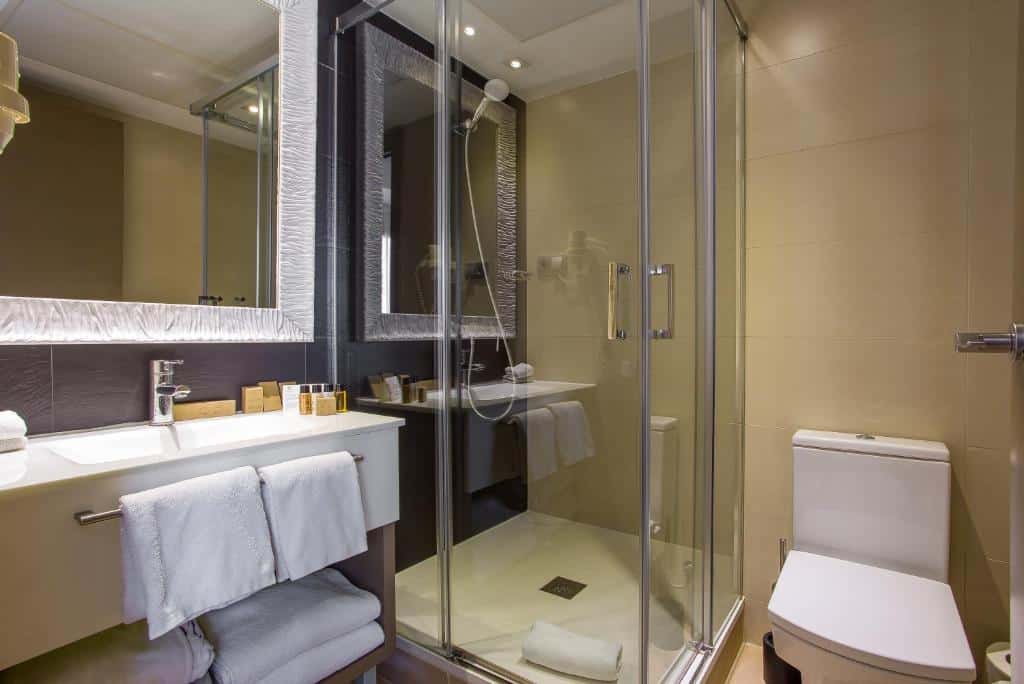Banheiro do Musik Boutique Hotel. A pia tem amenidades de banho, toalhas e espelho, e fica bem em frente ao box com chuveiro. A privada branca está ao lado da área do chuveiro.