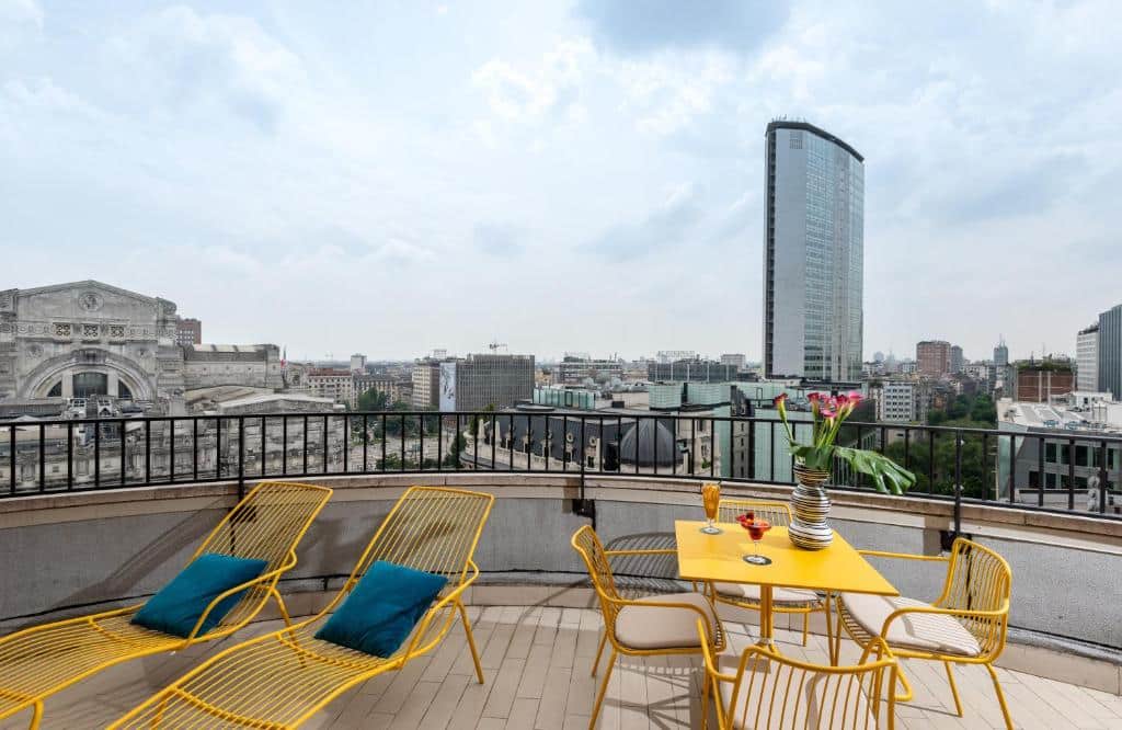 Varanda na cobertura do NYX Hotel Milan by Leonardo Hotels com vista para a estação central e outros prédios, na varanda há espreguiçadeiras, mesa e cadeiras de ferro amarelo