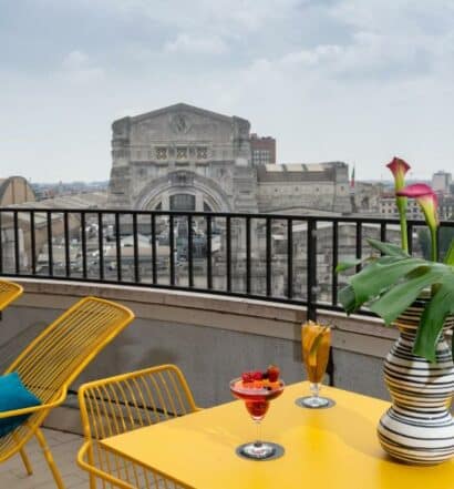 Varanda do NYX Hotel Milan by Leonardo Hotels com vista para a Estação Central, há uma mesa e cadeiras amarelas, sob a mesa, há um vaso de flor e duas taças com bebidas