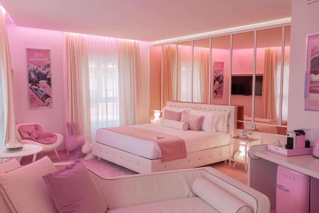 Quarto inteiro rosa do ODSweet Duomo Milano Hotel com cama de casal, cabeceira de espelho, uma poltrona e um sofá, há também uma pequena mesa com itens de café e um frigobar