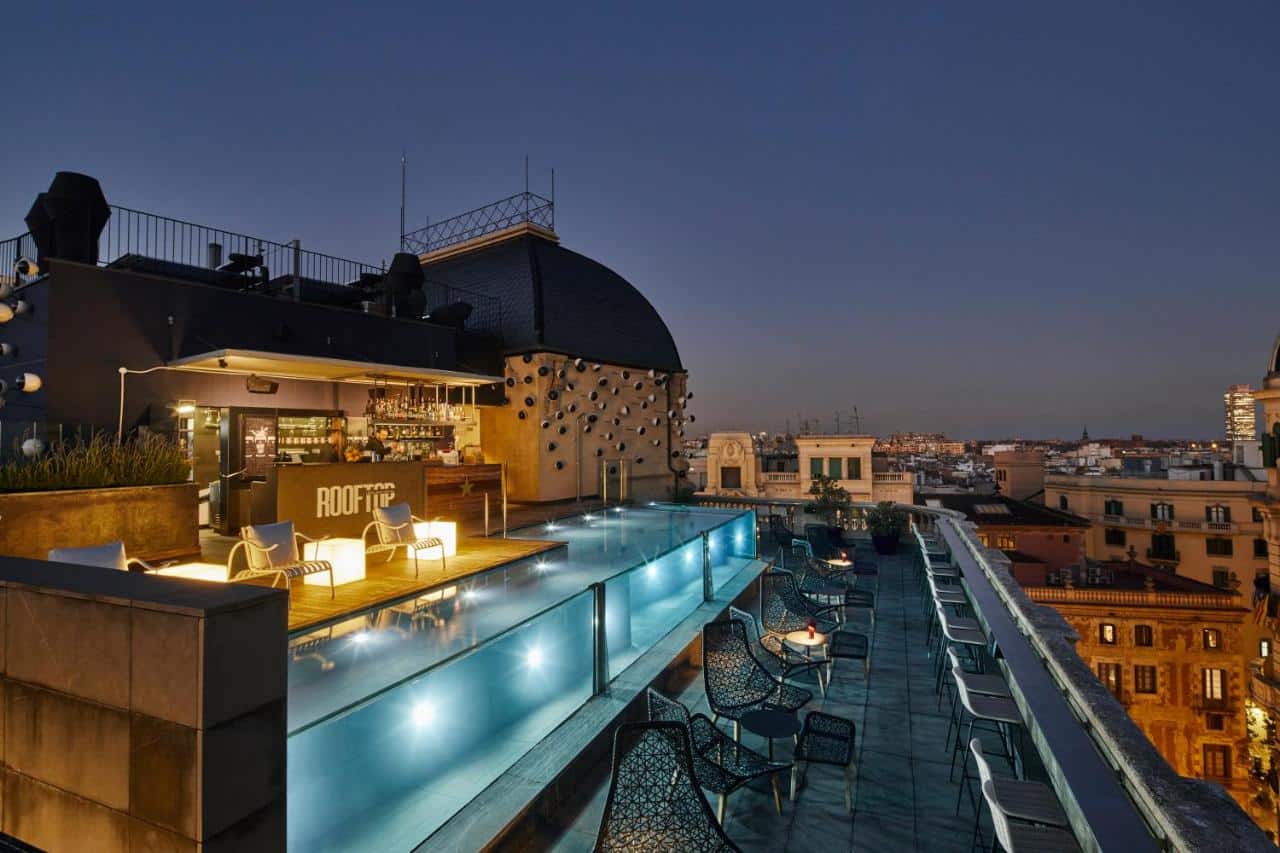 Terraço do Ohla Barcelona, uma das recomendações de hotéis boutique em Barcelona. A piscina com bordas de vidro está iluminada ao anoitecer, e cadeiras intercaladas com mesinhas encaram a vista da cidade.