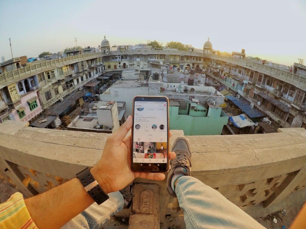 Pés de um homem, usando calça jeans e tênis, na sacada de um prédio, em old Delhi na india, segurando um celular com uma mão que tem um relógio preto no pulso. Ao fundo, tem a vista de um prédio com várias construções e casas juntas