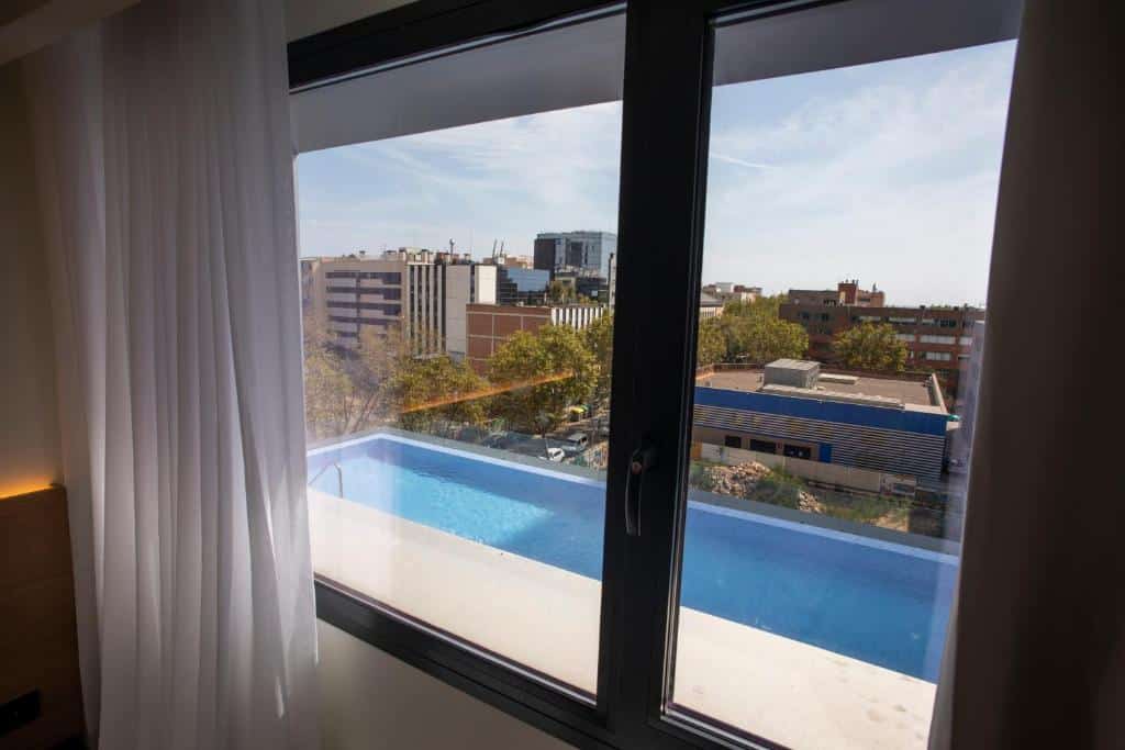 Vista da piscina num quarto do Hotel Paxton Barcelona. A janela de vidro tem cortinas brancas ao lado e proporciona vista para a piscina logo abaixo e a cidade de Barcelona ao longe. Há prédios e árvores ao redor do hotel.