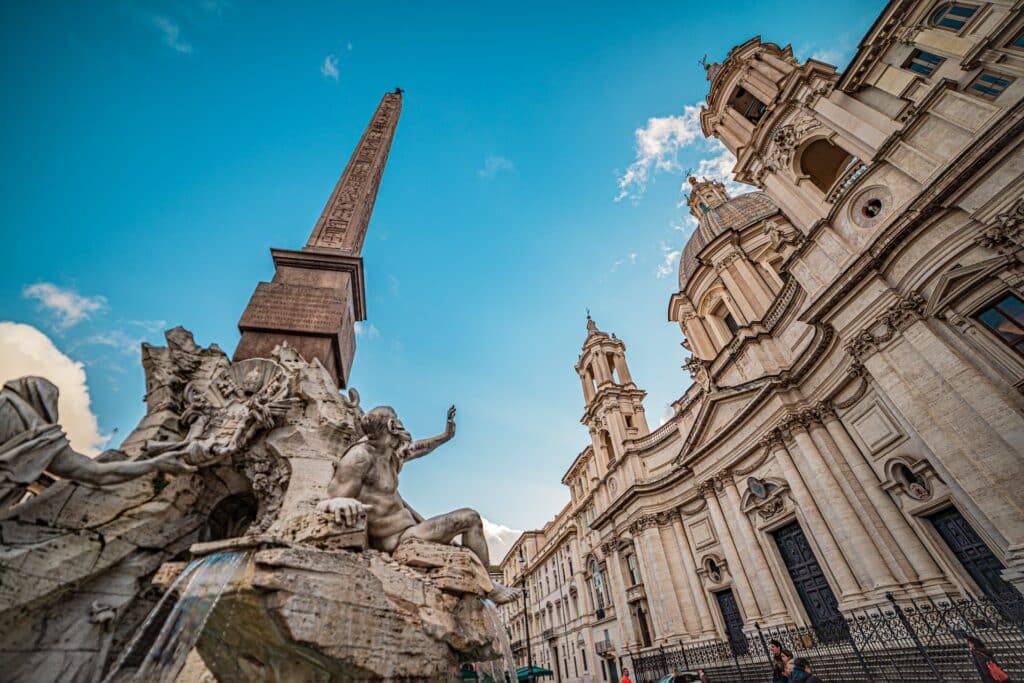 vista de baixo para cima da Piazza Navona com um obelisco, esculturas clássicas e um prédio também nesse estilo, super trabalhado em detalhes clássicos e fronte branca