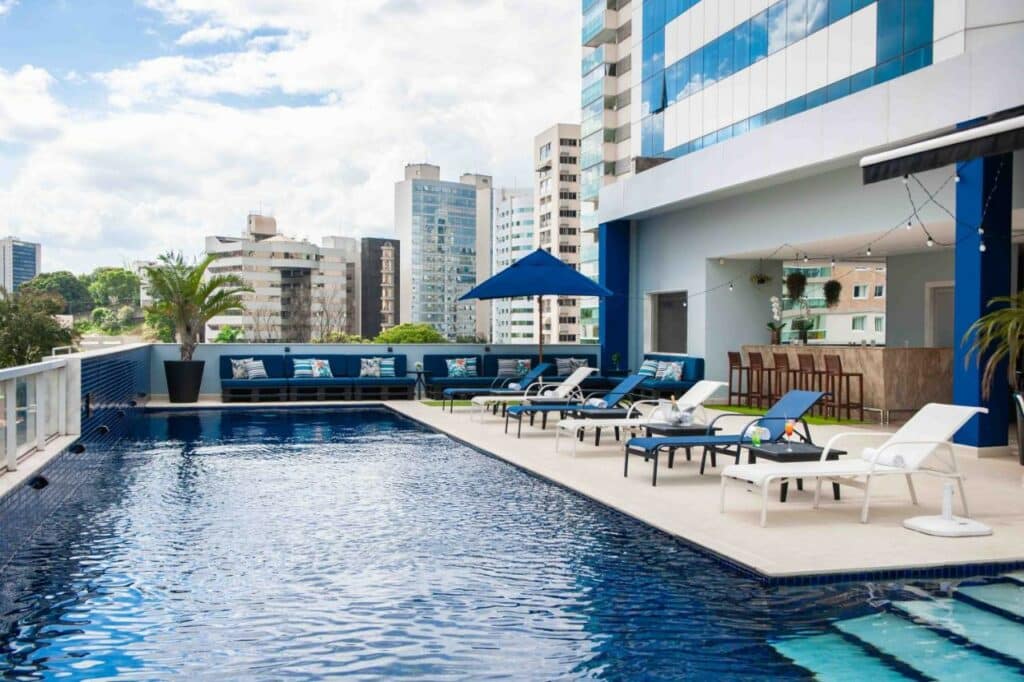 Grande piscina em uma varanda a céu aberto, rodeada por cadeiras de praia, guarda-sol, um bar e a cidade com vários prédios, na propriedade do Golden Tulip Porto Vitória.