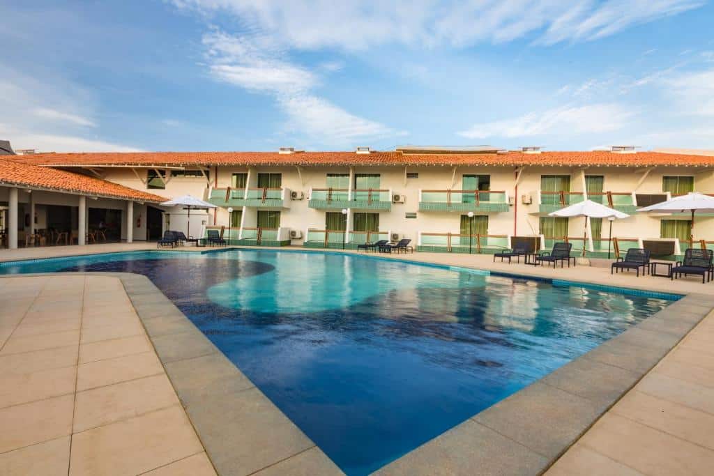Área externa de um hotel com piscina em frente as varandas dos quartos, espreguiçadeiras e guarda sol.
