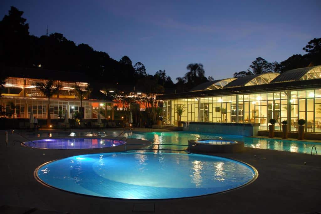Uma piscina grande e dividida em círculos, um de cada tamanho, no Villagio Embu. Atrás o hotel iluminado e com paredes de vidro. Imagem a noite. Foto para ilustrar post sobre Pousadas em Embu das Artes.