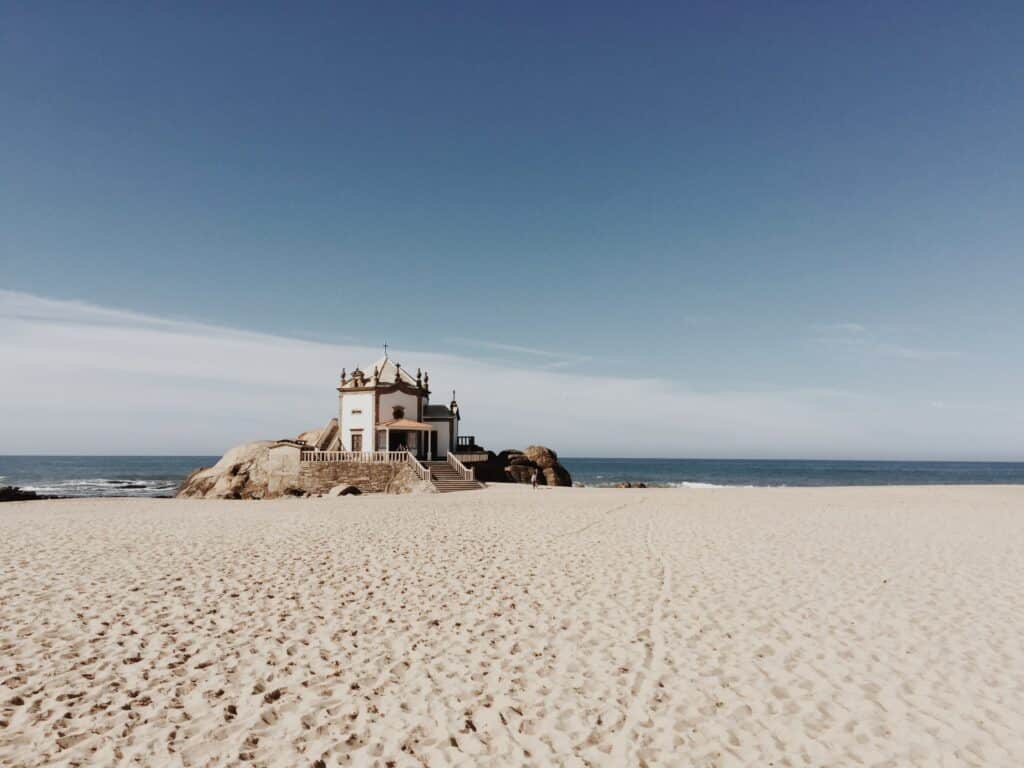 Praia Vila Nova de Gaia, durante o dia com areia a frente e ao fundo uma rocha com uma capela e ao fundo o mar.