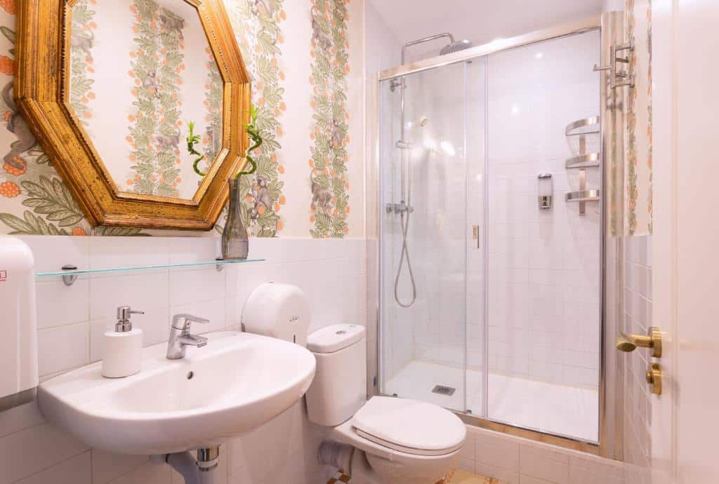 Banheiro do Primavera Hostel. A pia branca tem um espelho na parede acima e uma privada ao lado. O box de vidro com chuveiro e espaço para amenidades de banho está do outro lado da privada. As paredes do banheiro tem papel de parede florido.
