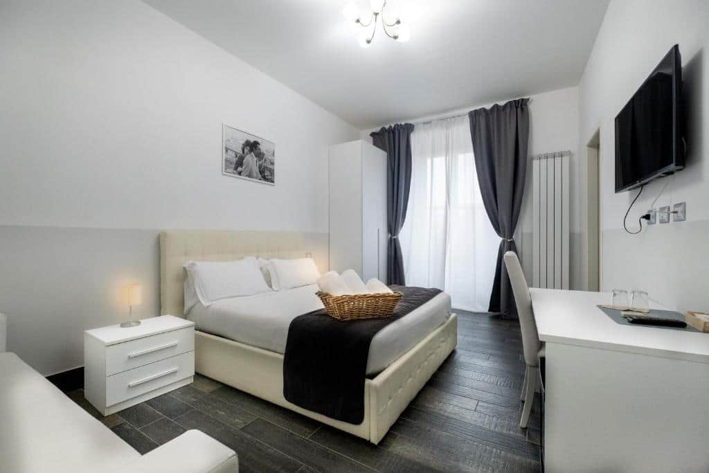 quarto do Arch Rome Suites, um dos hotéis no centro em Roma, com cama de casal grande, janela também bem grande com cortinas azuis escuras, em frente à cama, em que há mesinhas e luminária de ambos os lados, há uma mesa com cadeira e tv, existe ainda um sofá do outro lado