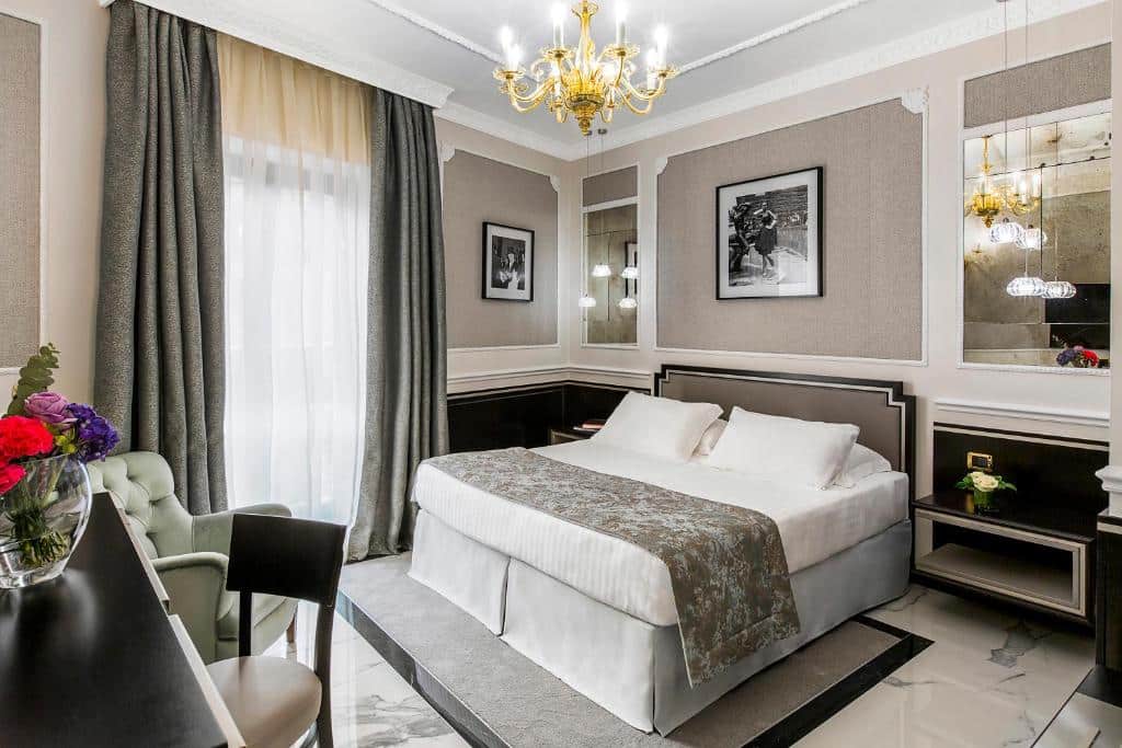 quarto do Baglioni Hotel Regina, um dos hotéis em Roma com um lustre dourado, cama enorme com mesinha e luminária de ambos os lados, mesa com tv acima e cadeira com banco acolchoado quadros na parede e janela grande com cortinas claras e escuras