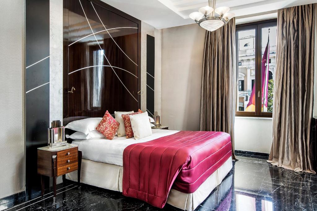 quarto do Baglioni Hotel Regina, um dos hotéis de luxo em Roma, com cama de casal, mesinha de madeira e luminária em ambos os lados, há um painel que combina com o piso feito em pedra preta contrastando com os detalhes em vermelho da cama, acima tem um lustre e as janelas são grandes