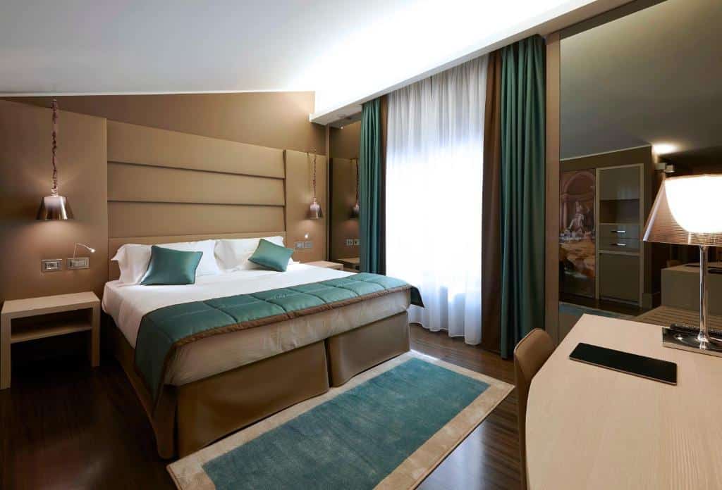quarto do Harry's Bar Trevi Hotel & Restaurant, um dos hotéis de luxo em Roma, com cama de casal, mesa, cadeira e tv em frente com detalhes em verde, há mesinha e luminária de ambos os lados com janela grande de um lado e espelho