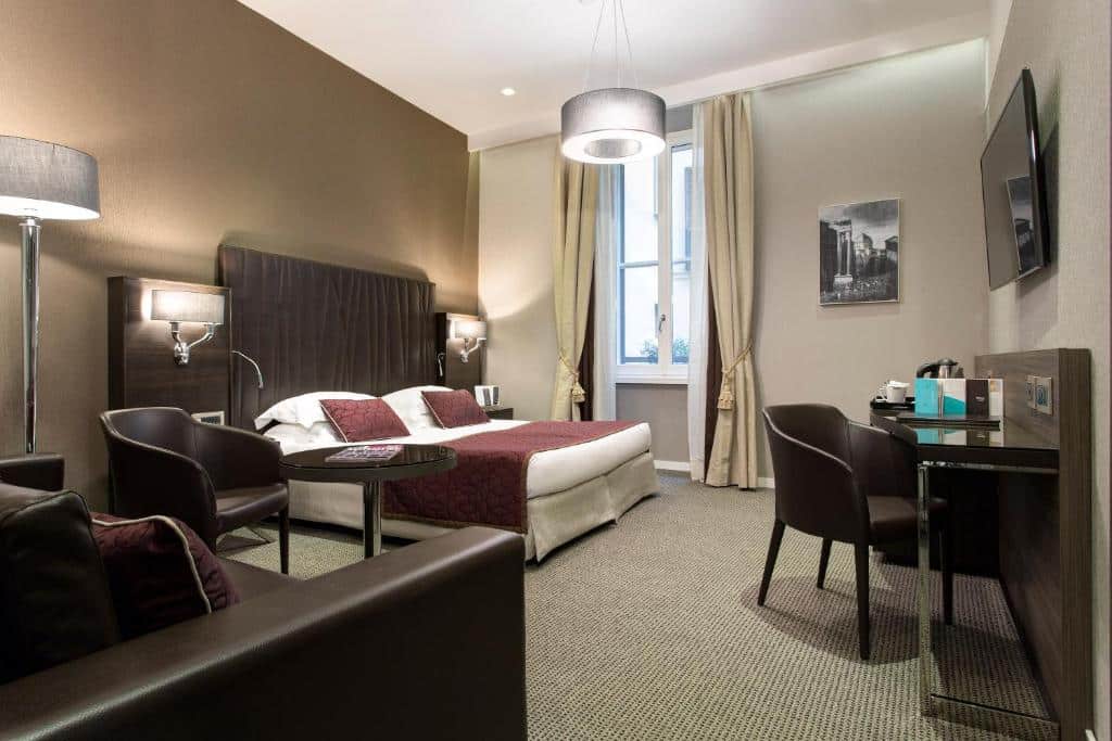 quarto amplo do Hotel Artemide, um dos hotéis de luxo em Roma, com cama de casal, mesinha e luminária de ambos os lados, lustre redondo, mesa com cadeiras em tons escuros e janela grande com cortinas claras