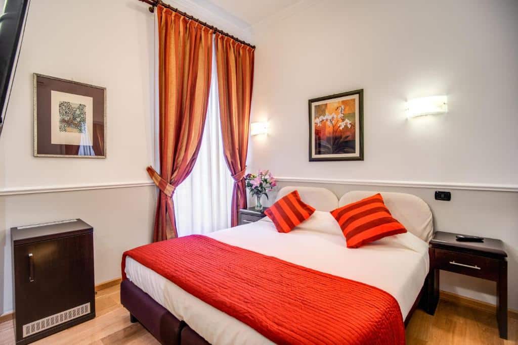 quarto do Hotel Everest Inn Rome, um dos hotéis baratos em Roma, com cama de casal em detalhes laranjas que combina com as cortinas na janela grande e há mesinhas em ambos os lados, há uma pequena cômoda também