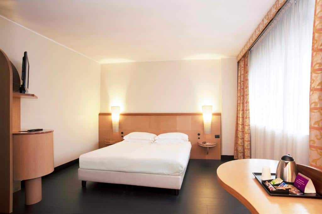 quarto do Mercure Roma West, com cama de casal, mesinha com luminária de ambos os lados, janela grande com cortinas, guarda-roupa e mais uma mesinha de madeira