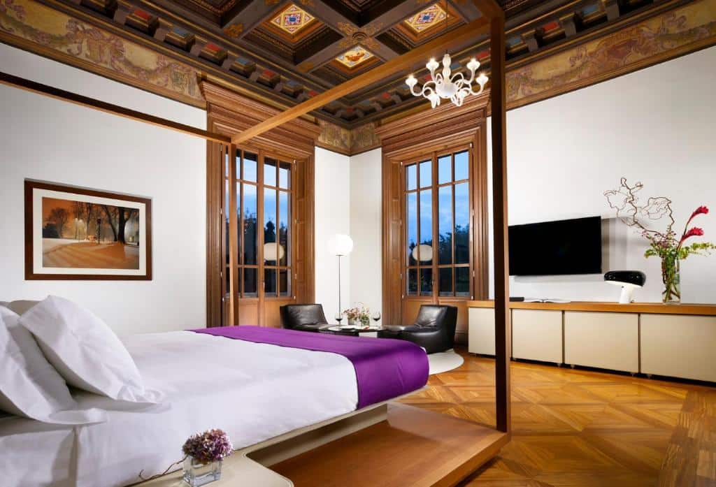 quarto do Palazzo Montemartini Rome, um dos hotéis de luxo em Roma, com cama de casal, janelas amplas e detalhes em madeira, mesinha dos lados com luminárias e cômoda acima de uma tv, com mesa e poltrona do outro lado