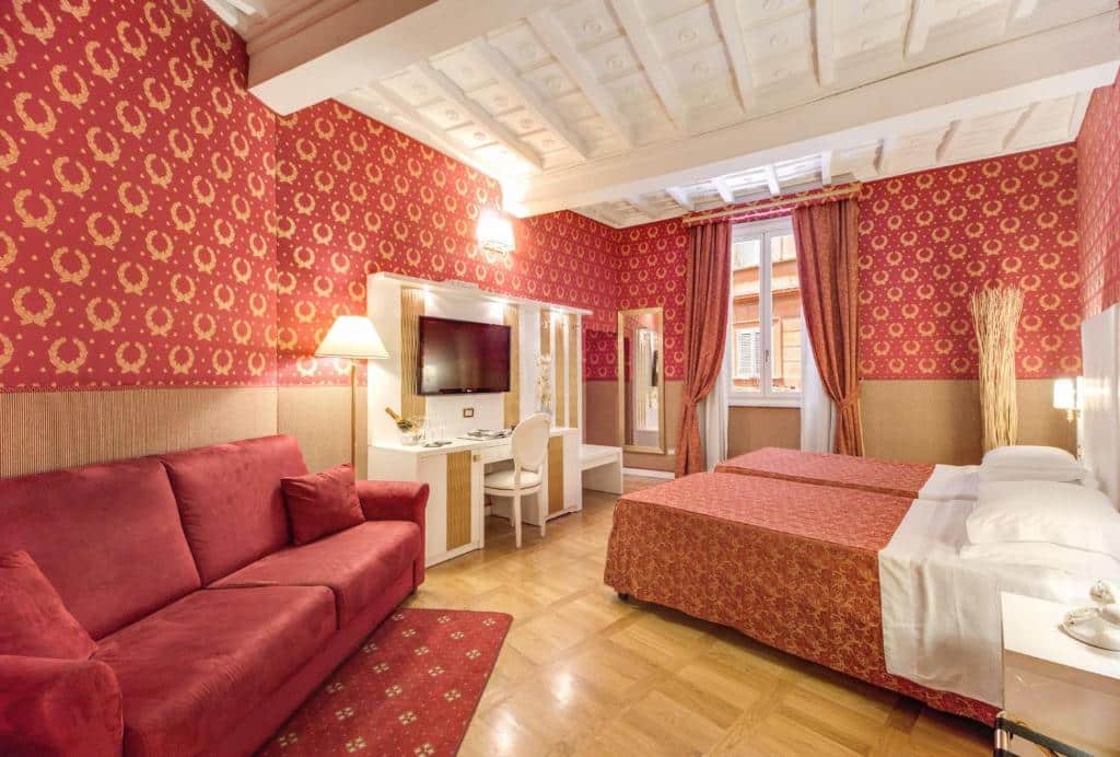 quarto do Relais Fontana Di Trevi Hotel, um dos hotéis no centro de Roma, com detalhes em vermelho, desde o papel de parede ao sofá e roupa de cama, há uma tv com balcão e cadeira em frente a duas camas que estão unidas, a janela é grande em madeira