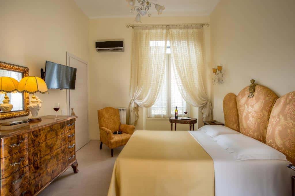 quarto do Roma Central Guest House, um hostel em Roma, com cama de casal com cabeceira em forma de concha, bem clássica, os móveis são de madeira, uma cômoda está À frente da cama com tv e do outro lado há uma janela grande com cortinas leves, uma mesa e poltronas amarelas, combinando com a roupa de cama