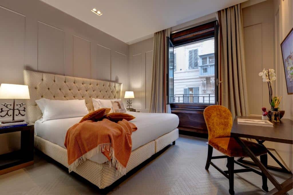 quarto do Singer Palace Hotel, um dos hotéis de luxo no centro de Roma, com cama de casal bem grande com manta e demais detalhes em laranja, há uma mesinha e luminária em ambos os lados, há uma cadeira e mesa de madeira em frente, com uma janela grande com cortinas