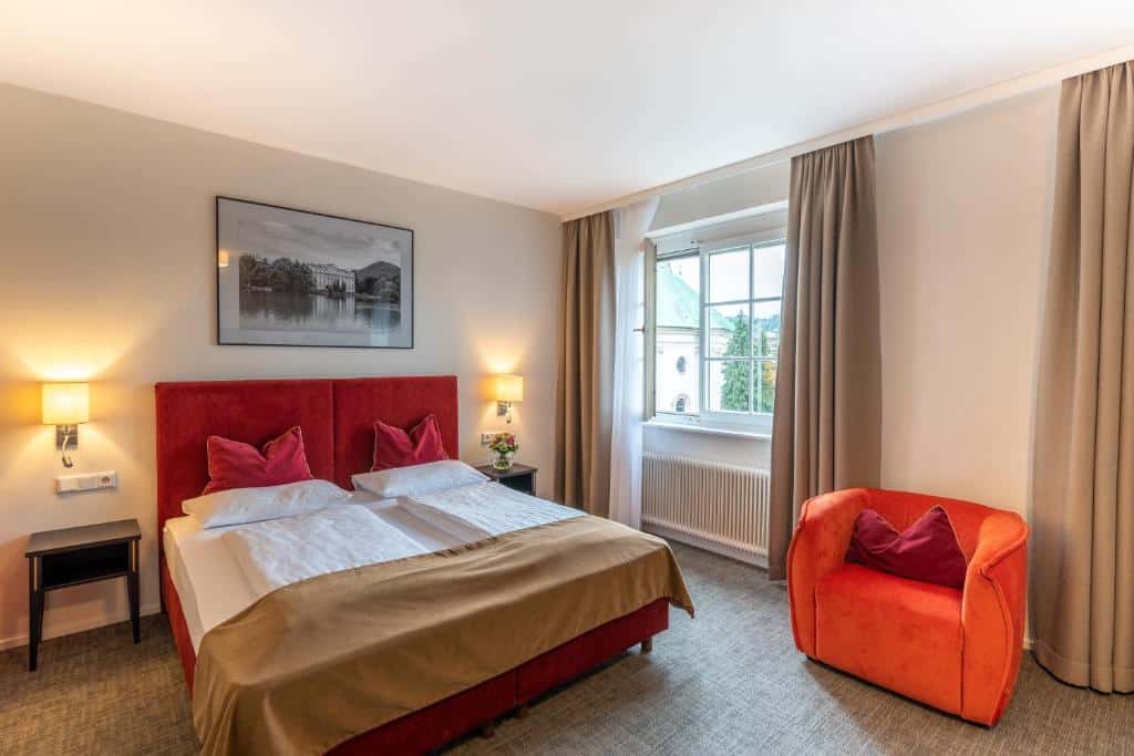 Quarto do hotel com uma cama de casal em tons de vermelho, branco e marrom, uma poltrona laranja, quadro na parede e janela de vidro com cortinas marrons.