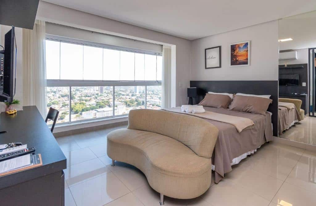 Quarto de apartamento com cama de casal, pequeno sofá a frente, espelho ao lado direito do quarto, mesa, cadeira e TV a frente da cama e janelas de vidro ao lado direito com vista para a cidade de Goiânia.