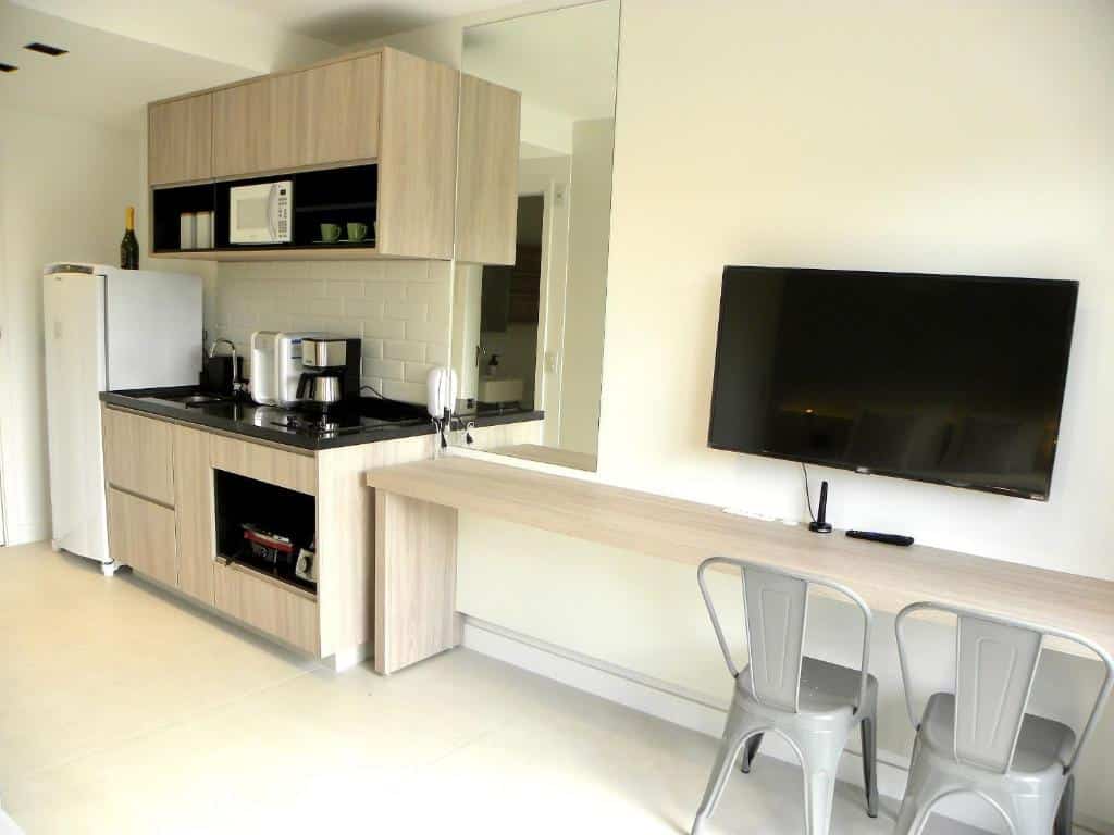 Uma mesa de escritório com duas cadeiras e uma televisão no lado direito. No lado esquerdo uma cozinha, armários, geladeira, forno, fogão e pia.