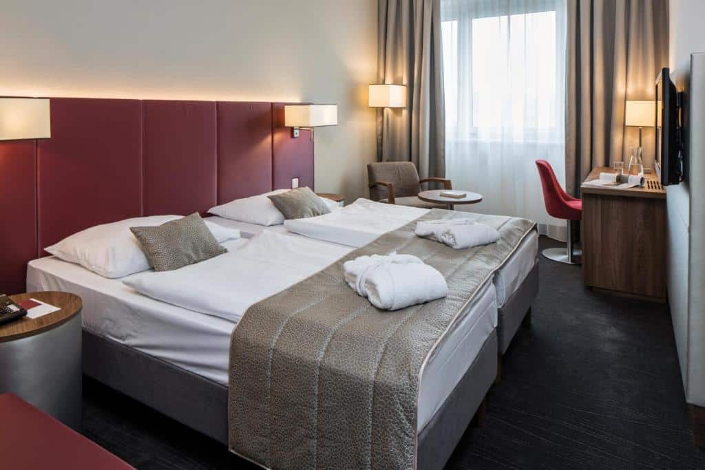 Quarto do hotel com uma cama de casal, parede branca com detalhes em tom de vinho, tv na parede, poltrona marrom e mesa com cadeira vermelha.