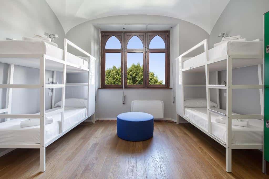 Quarto compartilhado do Babila Hostel & Bistrot com duas beliches brancas, há uma janela no centro do quarto e um bufê para sentar-se