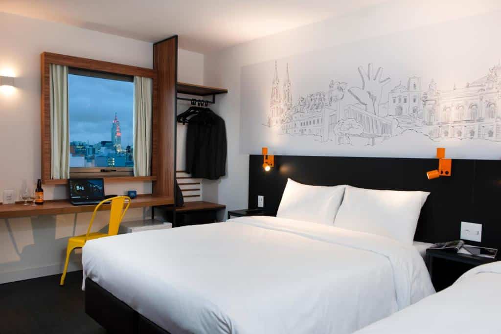 Quarto do hotel com uma cama de casal branca, parede preta e branca com desenhos, cabides com roupas, mesa de madeira com cadeira amarela e janela.