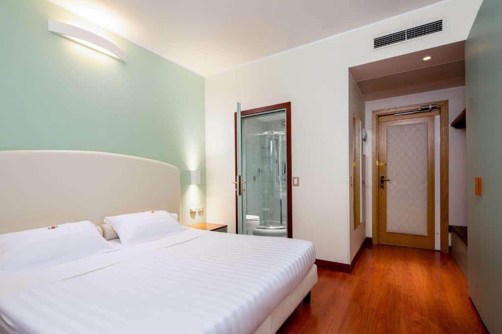 Quarto do Best Western Air Hotel Linate com uma cama de casal, um armário de conceito aberto, um espelho de corpo inteiro ao lado da porta de entrada, e uma pequena mesinha de cabeceira com uma luminária presa na parede