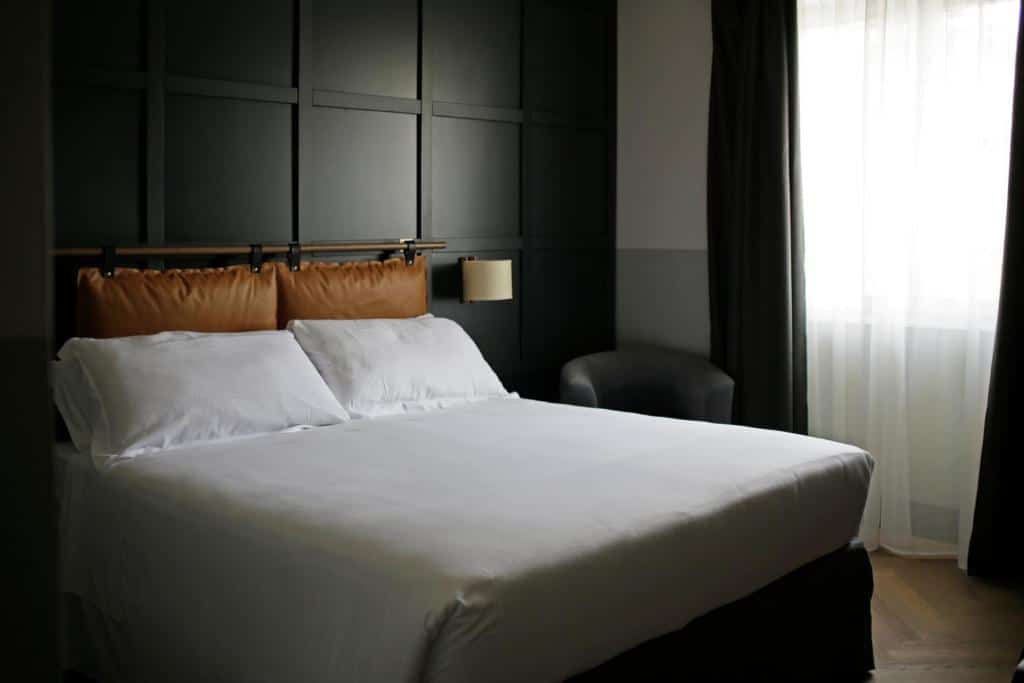 quarto do c-hotels Club House Roma, um dos hotéis baratos em Roma, com cama de casal grande, mesinha com luminária em ambos os lados, a parede tem textura, há uma poltrona ao lado e uma janela grande com cortinas escuras