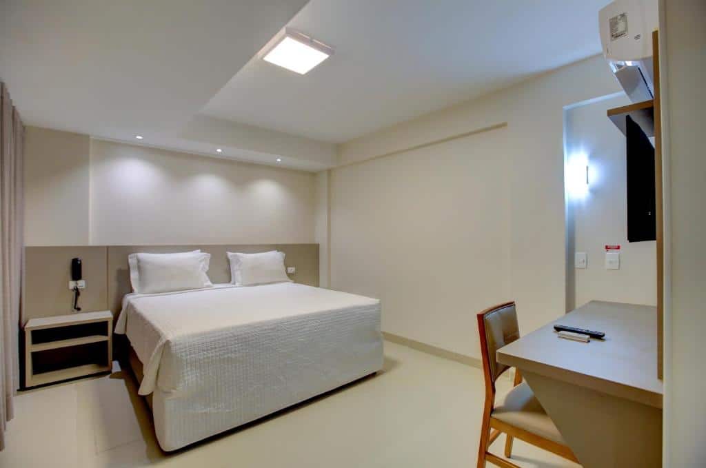 Quarto com cama de casal com lençóis brancos, paredes em tons de bege, móveis de madeira e cadeira a frente de uma mesa. Imagem para ilustrar o post hotéis em Goiânia.