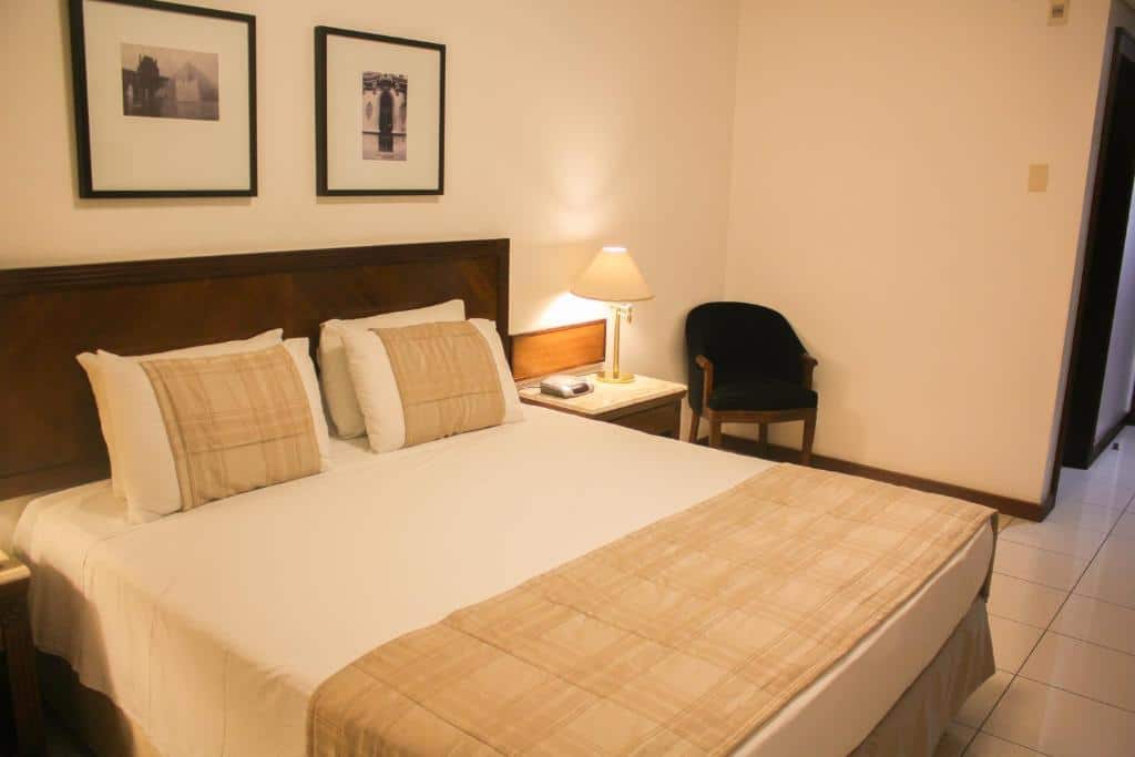 Quarto de hotel com paredes brancas, quadros decorativos, poltrona preta, abajur, cama de casal e iluminação amarelada. Imagem para ilustrar o post hotéis em Teresina.
