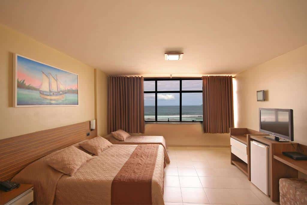 Quarto de hotel espaçoso com uma casa de casal, uma cama de solteiro, quadro decorativo, cômoda com frigobar, televisão e grande janela com cortinas e vista para o mar. Imagem para ilustrar o post hotéis em Cabo Frio.