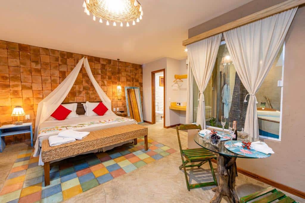 Vista do quarto do hotel Vila Parnaiba com grande cama de casal, mesa, cadeiras e cortinas brancas.
