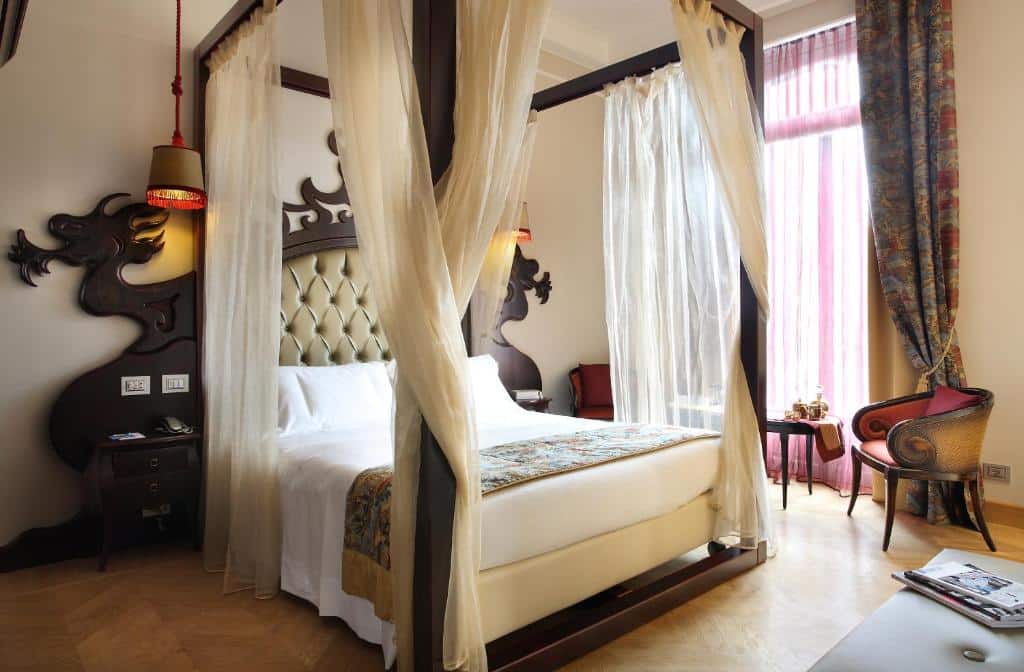Quarto do Château Monfort - Relais & Châteaux com uma cama de casal ampla com uma espécie de bangalô com panos jogados nos arredores dela, há também uma janela, uma pequena mesa com duas poltronas, e luminárias presas no teto