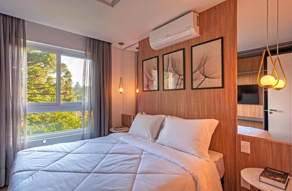 quarto do Cityhome Bav Residenz Gramado com uma cama de casal, um painel de madeira com cabeceira, dois espelhos grandes em cada lado da cama e uma janela ampla, no lado esquerdo da foto, mostrando as árvores araucárias da região.