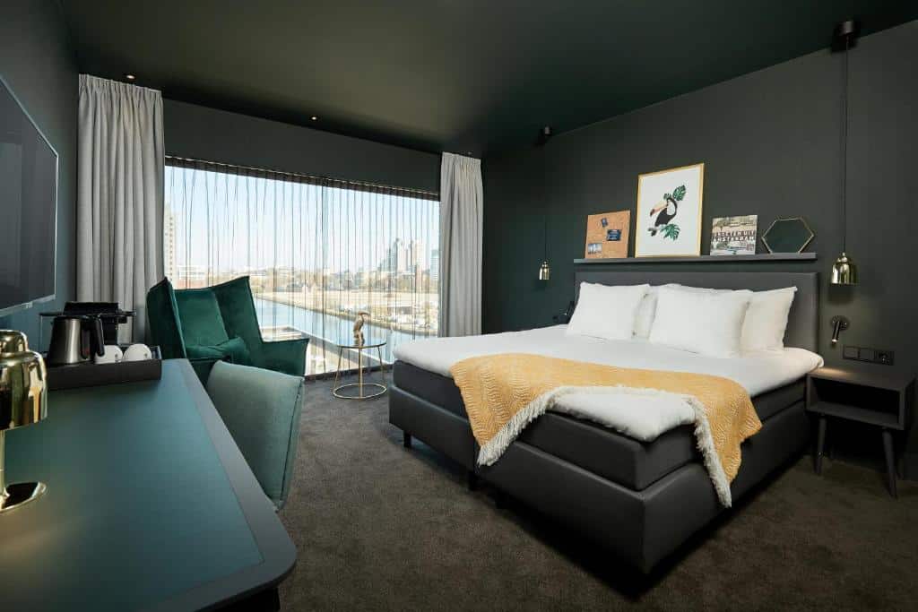 Quarto Comfort do Van der Valk Hotel, de 31 m², com cama de casal, poltrona verde escuro, e uma mesa com cadeira. Há uma janela panorâmica com vista da cidade, e o chão é de carpete