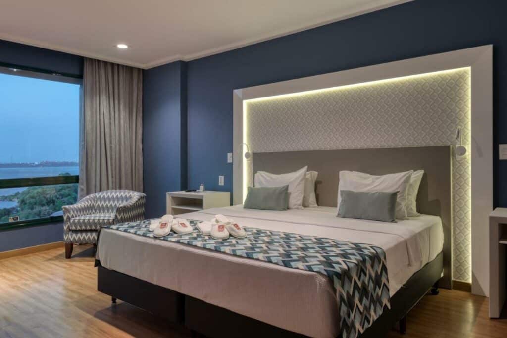 Quarto no Comfort Suítes Vitória com uma cama de casal, cabeceira iluminada, mesinha, poltrona e uma janela com cortina e vista para a cidade, para representar hotéis em Vitória.