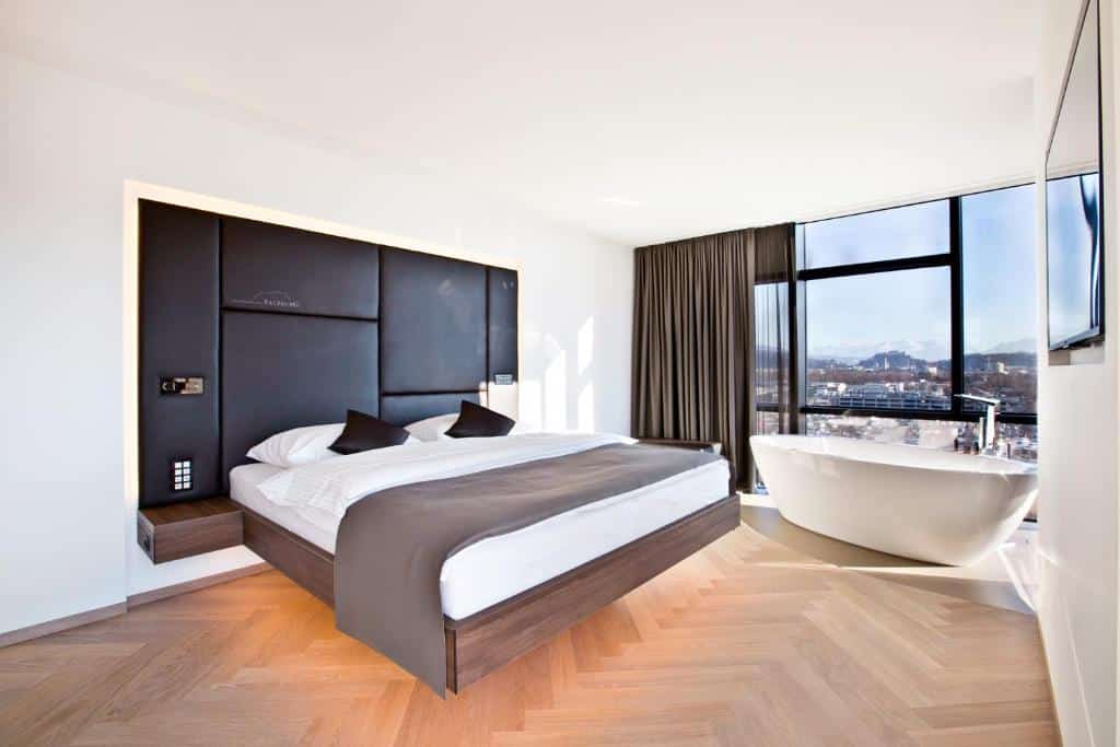 Quarto do hotel com cama suspensa, chão de madeira, banheira branca e uma parede de vidro com vista para a paisagem e uma cortina para tampar a visão.