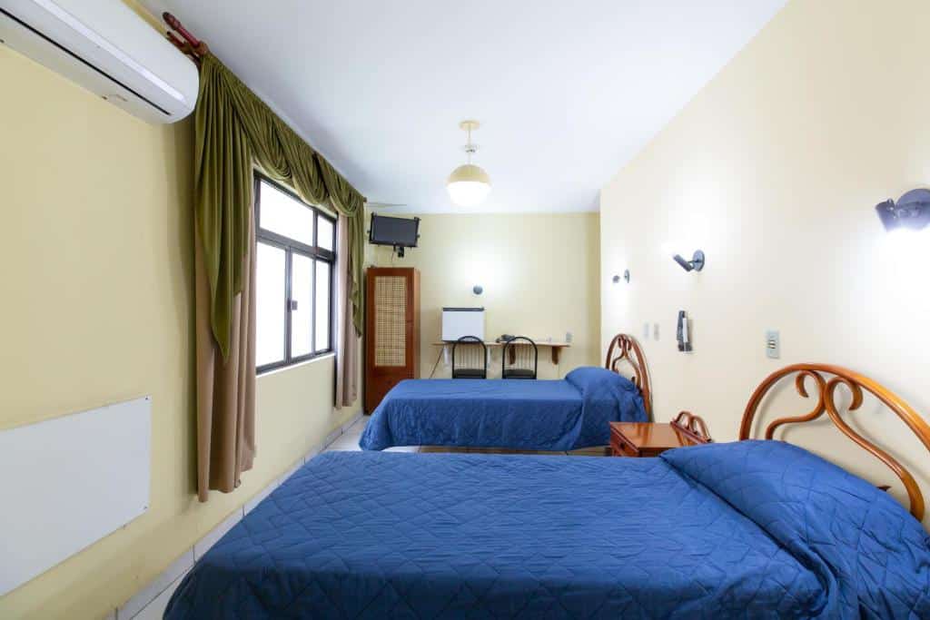 Quarto do hotel com duas camas azuis, uma mesinha separando as duas, ao fundo uma mesa com cadeiras, uma tv, um armário e uma janela de vidro com cortina verde.
