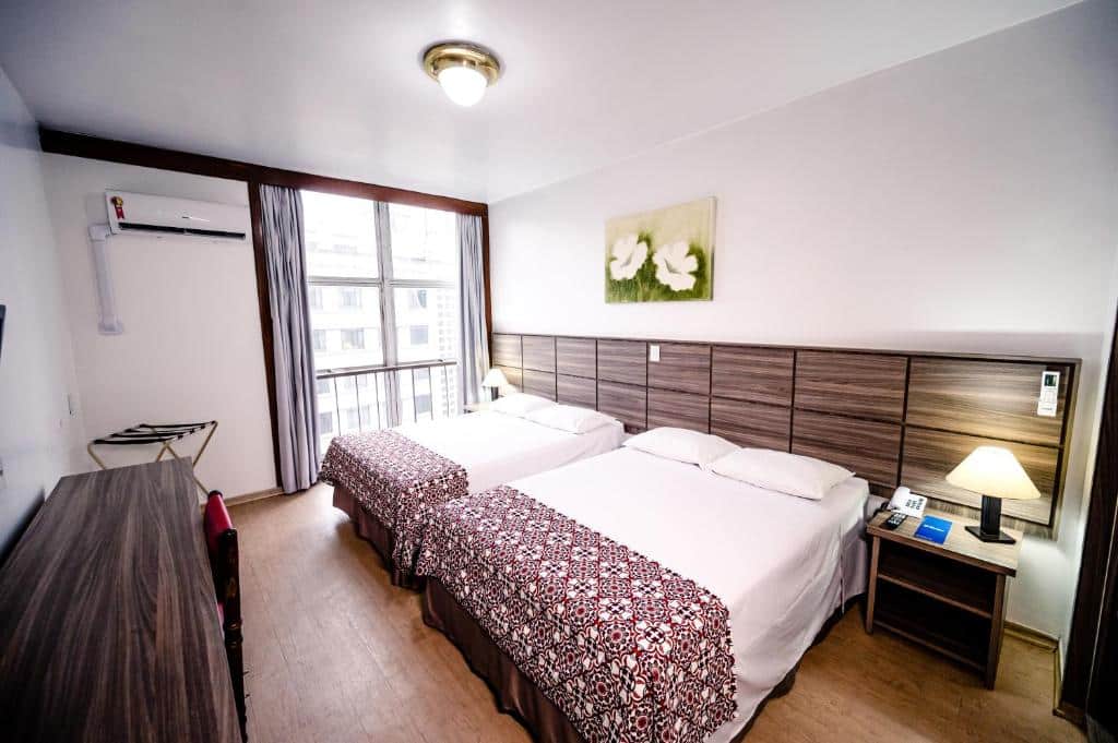 Quarto do hotel com duas camas, móveis de madeira, quadro na parede branca, ar condicionado e varanda com janelas de vidro.