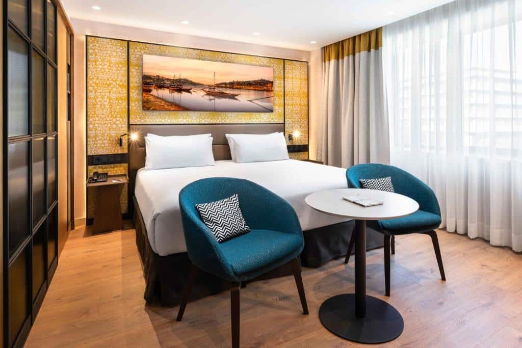 Quarto do Eurostars Aliados com cama de casal do lado esquerdo, duas cadeiras azuis no pé da cama com uma mesa redonda. Representa hotéis de luxo no Porto.